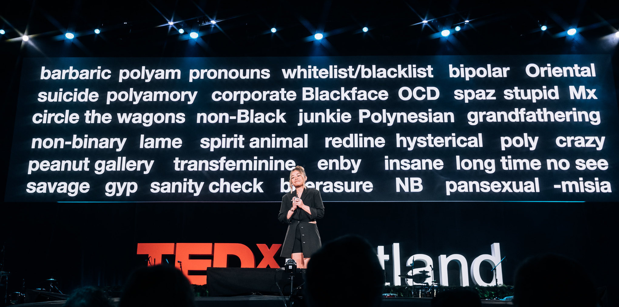 Tatiana Mac, smiling, cross-legged, standing in black suit on a TEDxPortland stage with giant words from Self-Defined app projected behind them